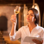 Professional winemaker female tasting a glass of white wine in his traditional cellar surrounded by wooden barrels. She uses a digital tablet