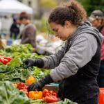 A young woman dressed in a gray sweatshirt and black gloves carefully selects vibrant peppers and leafy greens at a bustling farmers market. Surrounding her are various stalls filled with fresh produce, showcasing the local agricultural bounty during the autumn season.